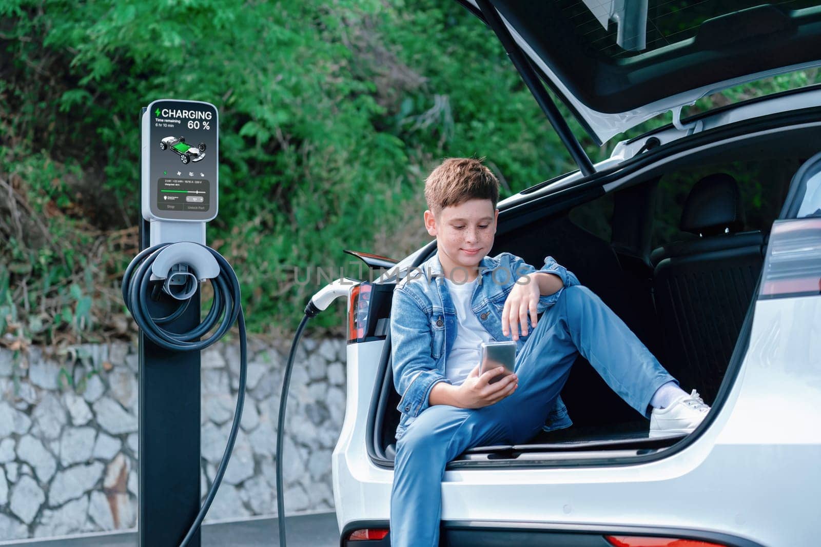 Little boy sitting on car trunk, using smartphone while recharging eco-friendly car from EV charging station. EV car road trip travel as alternative vehicle using sustainable energy concept. Perpetual