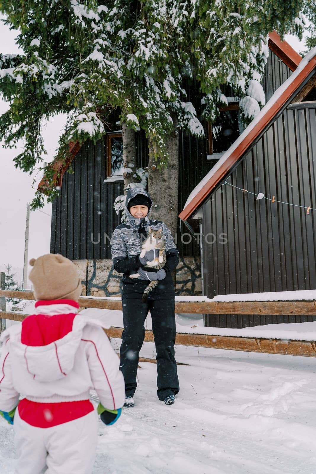 Small child looks at his mother with a tabby cat in her arms standing at the fence of a snow-covered house. Back view by Nadtochiy