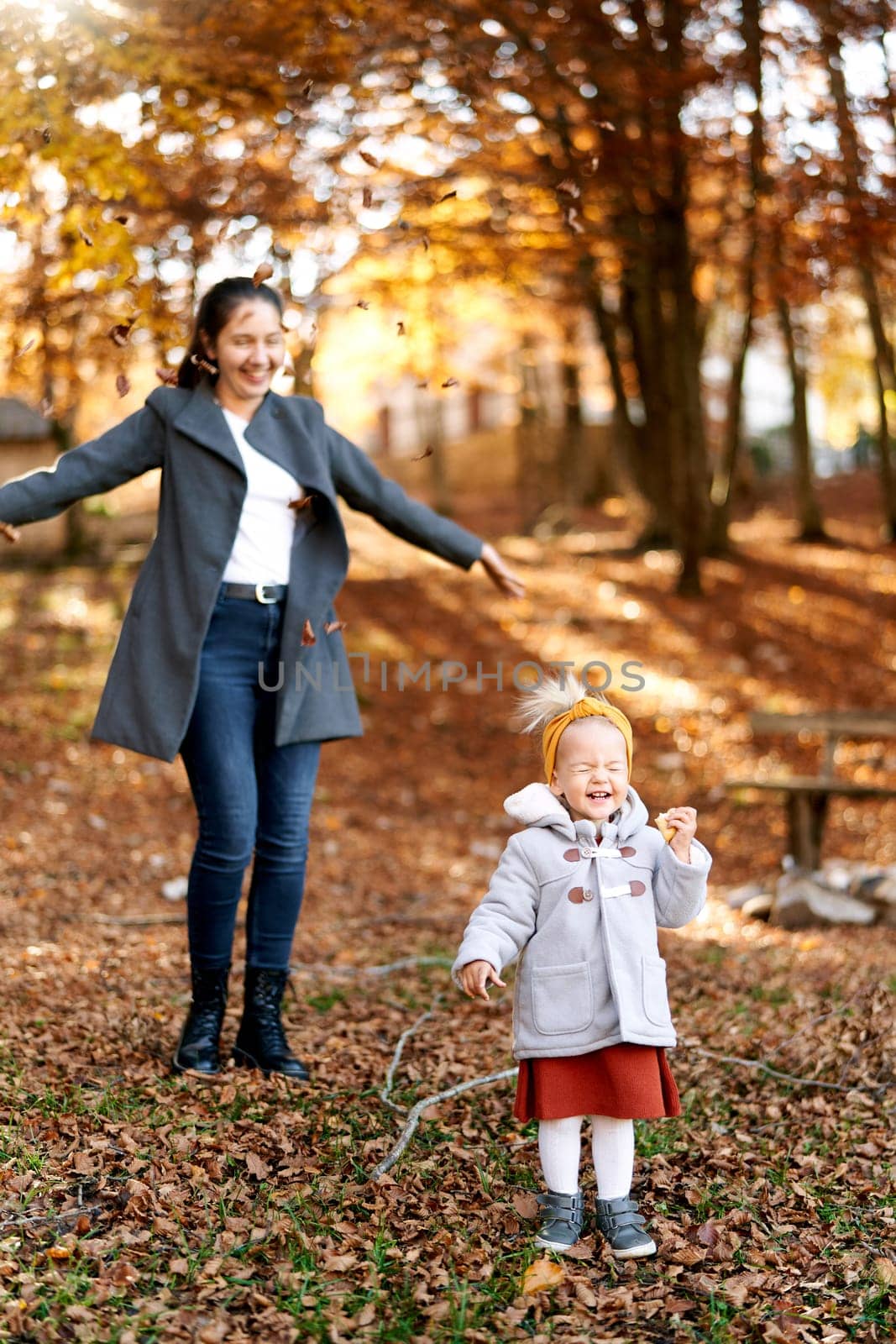Smiling mom throws up fallen leaves over a little laughing girl in an autumn park by Nadtochiy