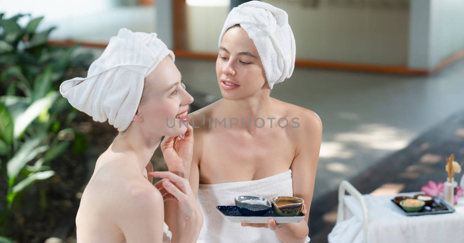 A portrait of beautiful woman in white towel receiving herbal facial mask by her friend at outdoor. Attractive spa girl putting face mask on pretty female with beautiful skin. Tranquility.