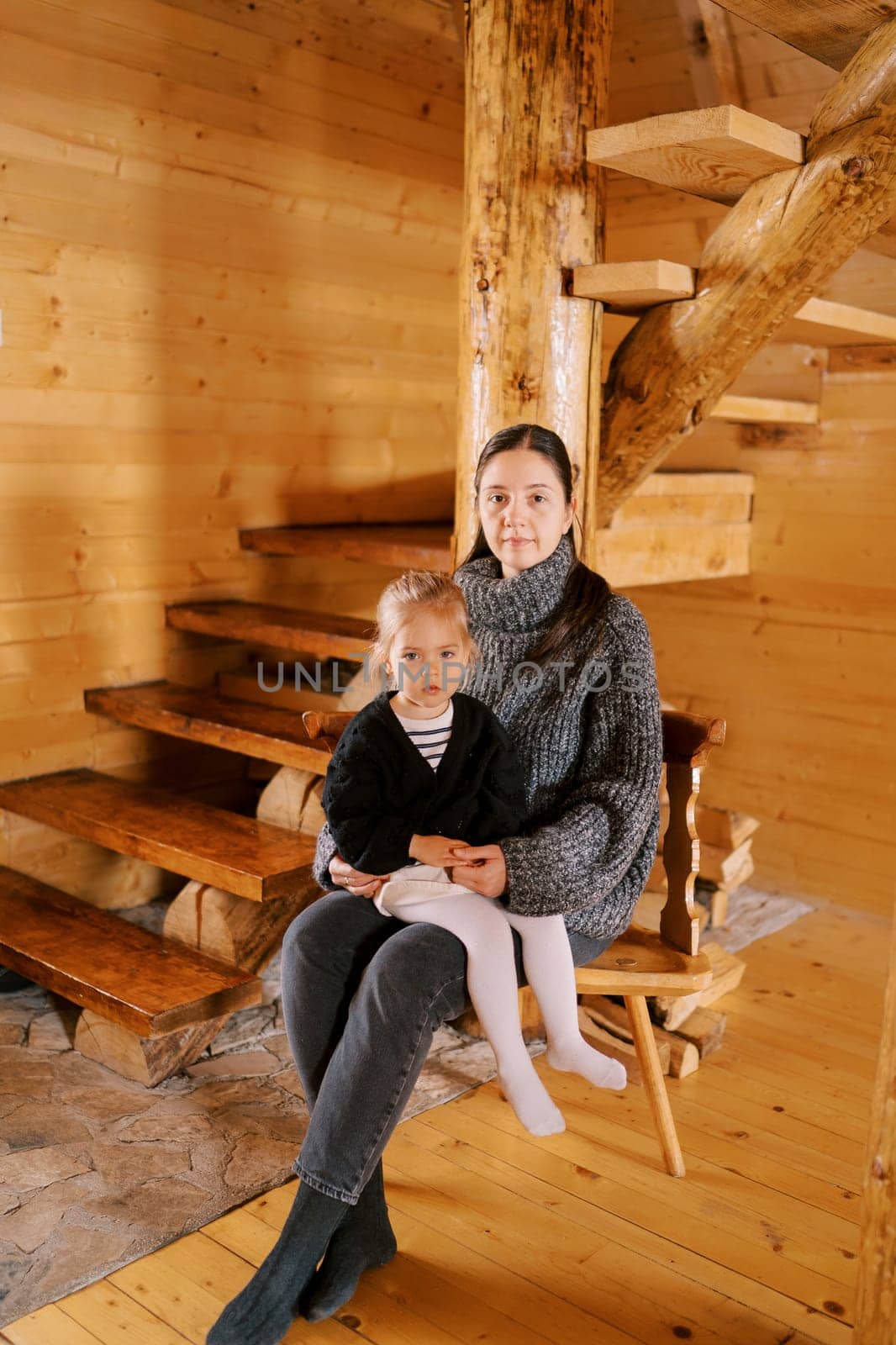Mother with a little girl on her lap sits on a chair in a wooden chalet near the stairs. High quality photo