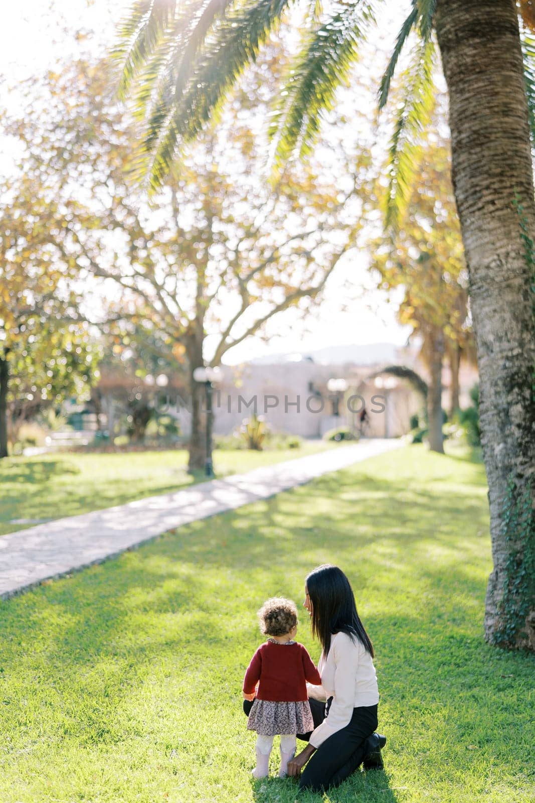 Mom squatted down next to the little girl near the tree. Back view by Nadtochiy