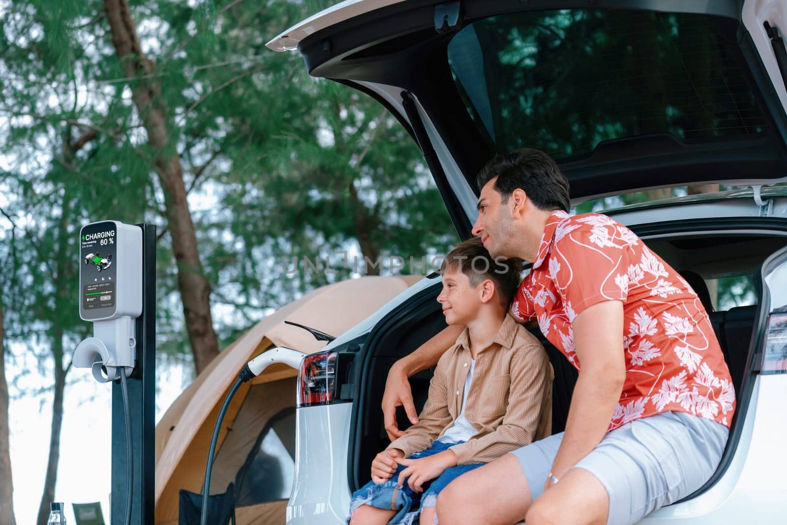 Outdoor adventure and family vacation camping in nature travel by eco friendly car for sustainable future. Father and son sit on trunk, charging EV car with EV charging station in campsite. Perpetual