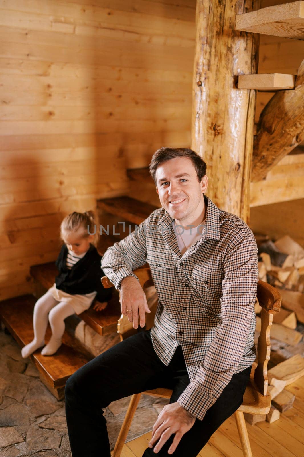 Smiling dad sitting on chair next to little girl sitting on stairs in wooden house. High quality photo