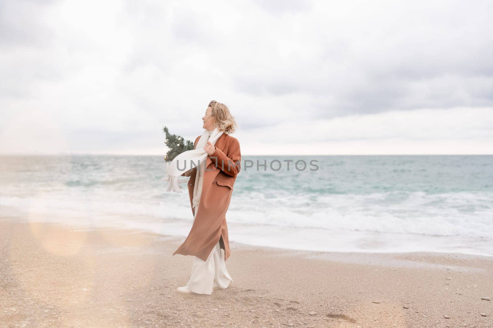 Blond woman Christmas tree sea. Christmas portrait of a happy woman walking along the beach and holding a Christmas tree in her hands. She is wearing a brown coat and a white suit. by Matiunina