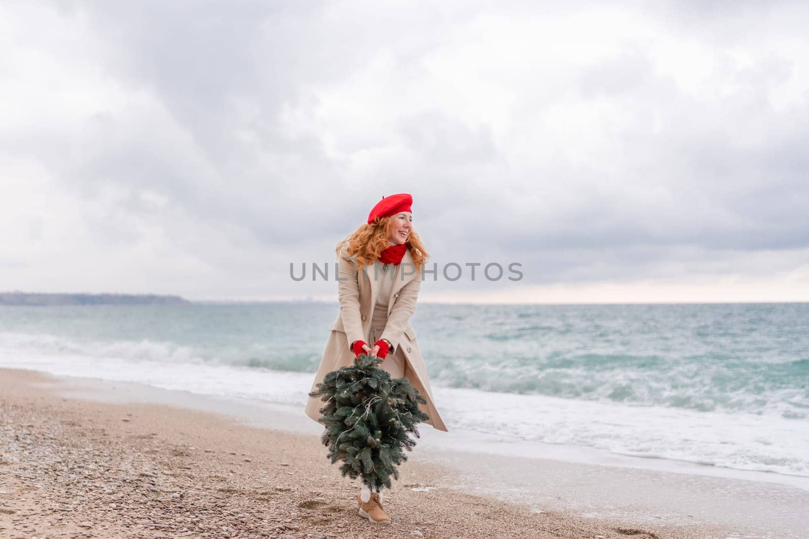 Redhead woman Christmas tree sea. Christmas portrait of a happy redhead woman walking along the beach and holding a Christmas tree in her hands. She is dressed in a light coat and a red beret. by Matiunina