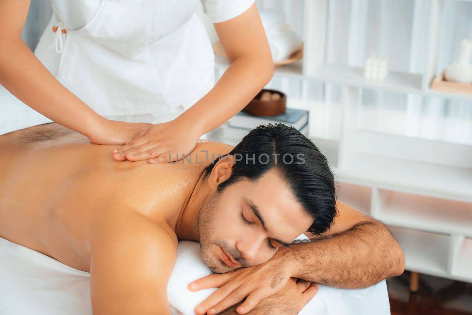 Caucasian man customer enjoying relaxing anti-stress spa massage and pampering with beauty skin recreation leisure in day light ambient salon spa at luxury resort or hotel. Quiescent