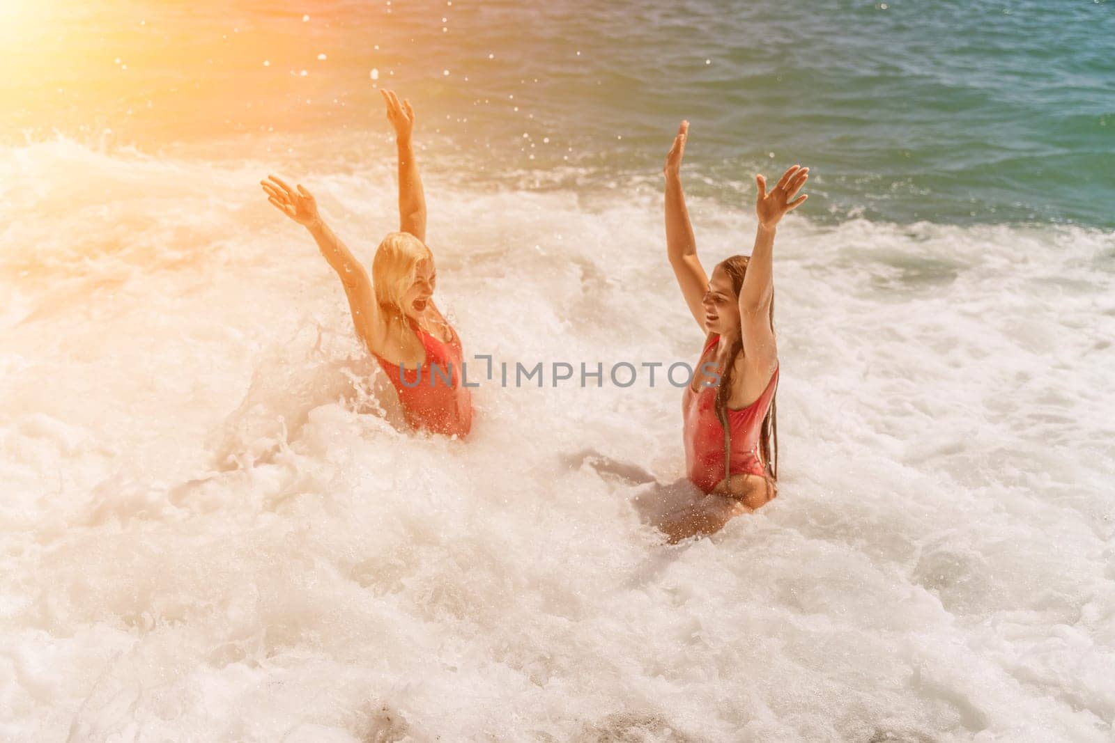 Women ocean play. Seaside, beach daytime, enjoying beach fun. Two women in red swimsuits enjoying themselves in the ocean waves and raising their hands up. by Matiunina