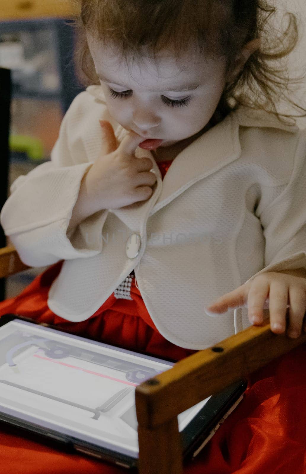 Portrait of one little Caucasian baby girl holding her finger in her mouth and looking down at a tablet with a cartoon, sitting on a wooden children's chair, close-up side view.