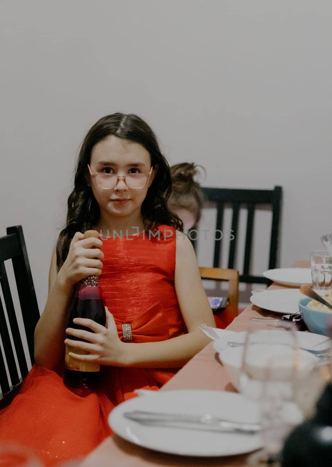 Portrait of one young beautiful Caucasian brunette girl in a red dress sitting at a table and holding a bottle of non-alcoholic champagne for children in her hands, close-up side view.