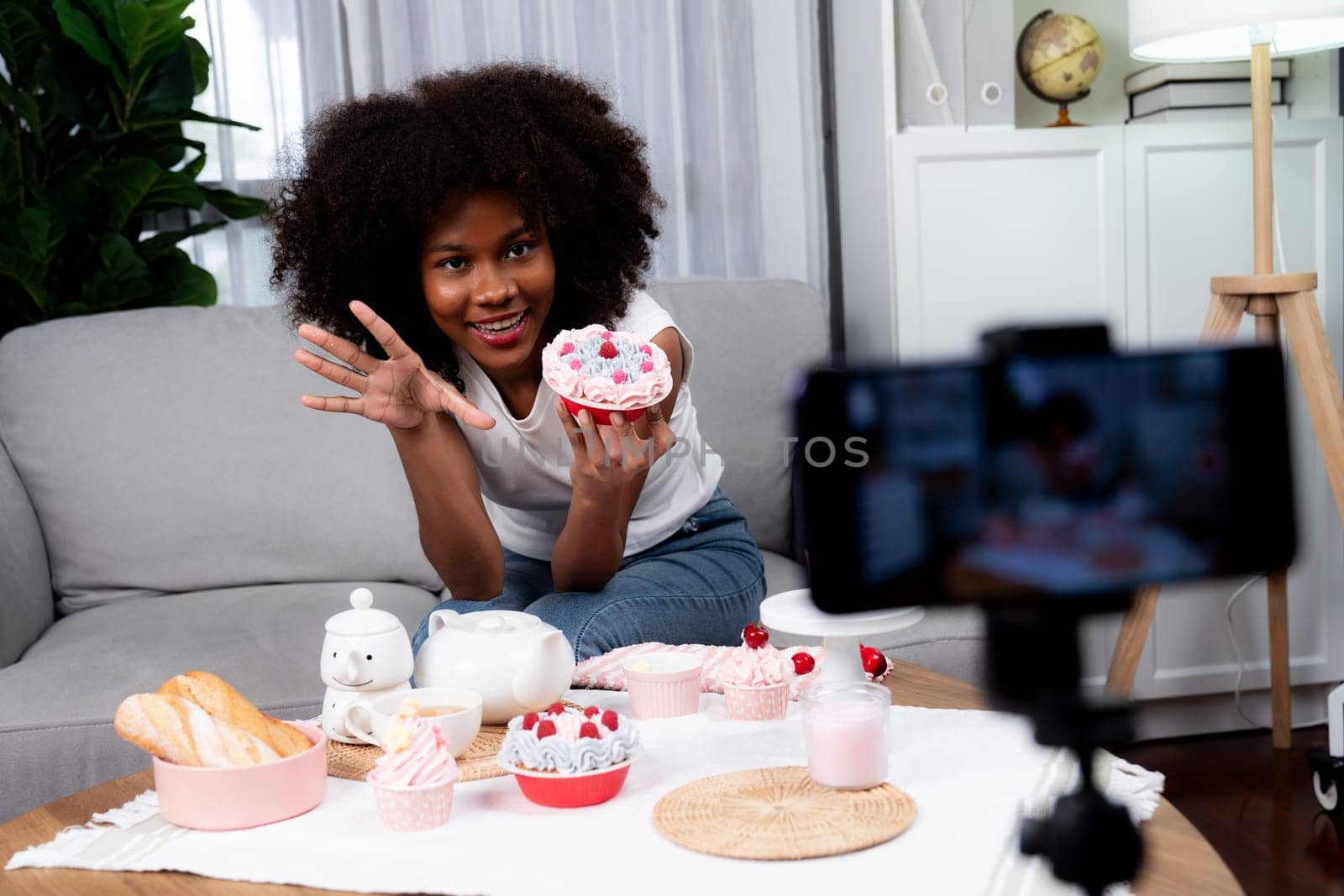 Beautiful young African blogger presenting piece of cupcake in concept special cuisine with video record on smartphone. Content creating of social media with favorite sweets bakery dish. Tastemaker.