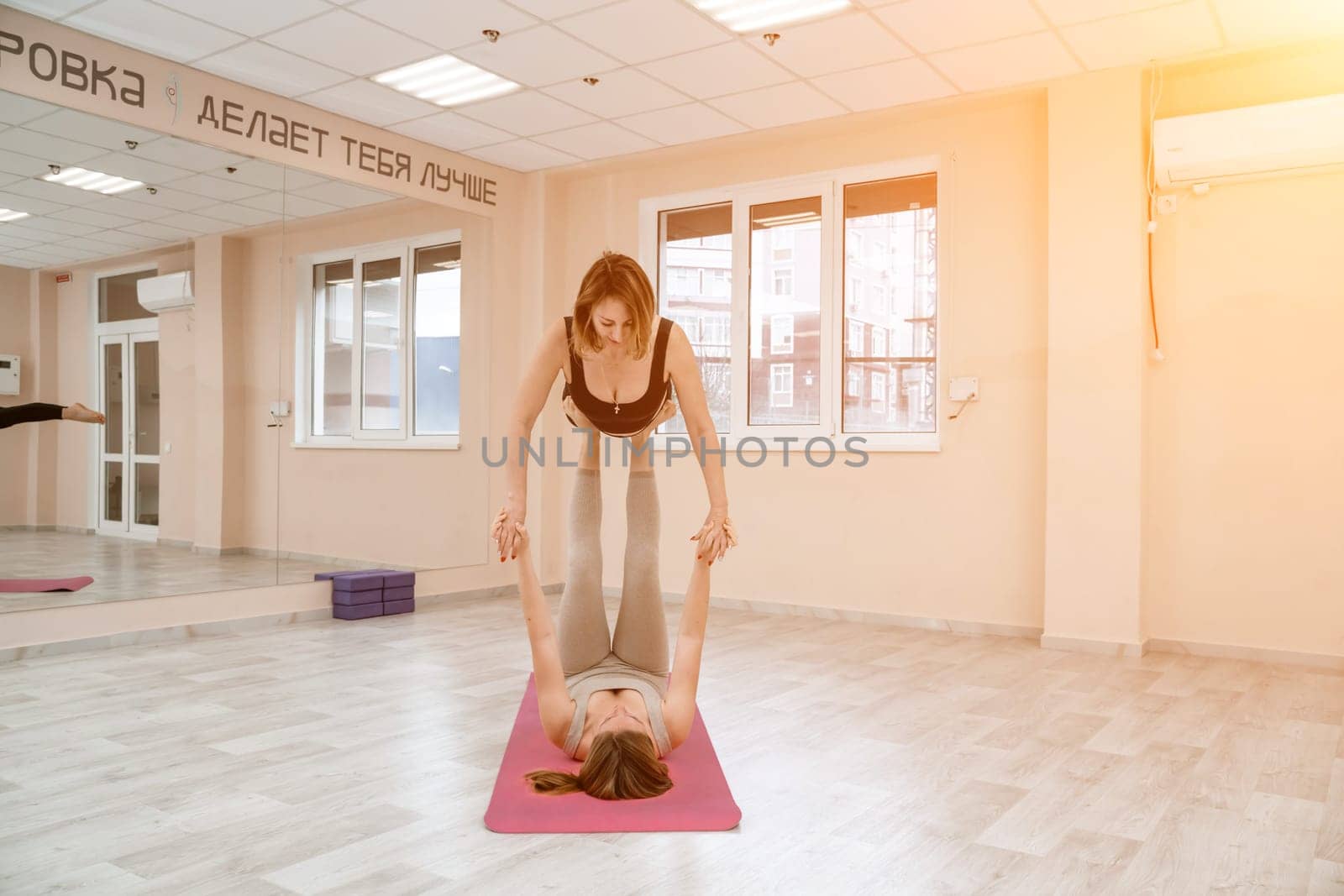 Two women do acroyoga on a light floor near a large mirror in a yoga studio. They are wearing leggings and tops