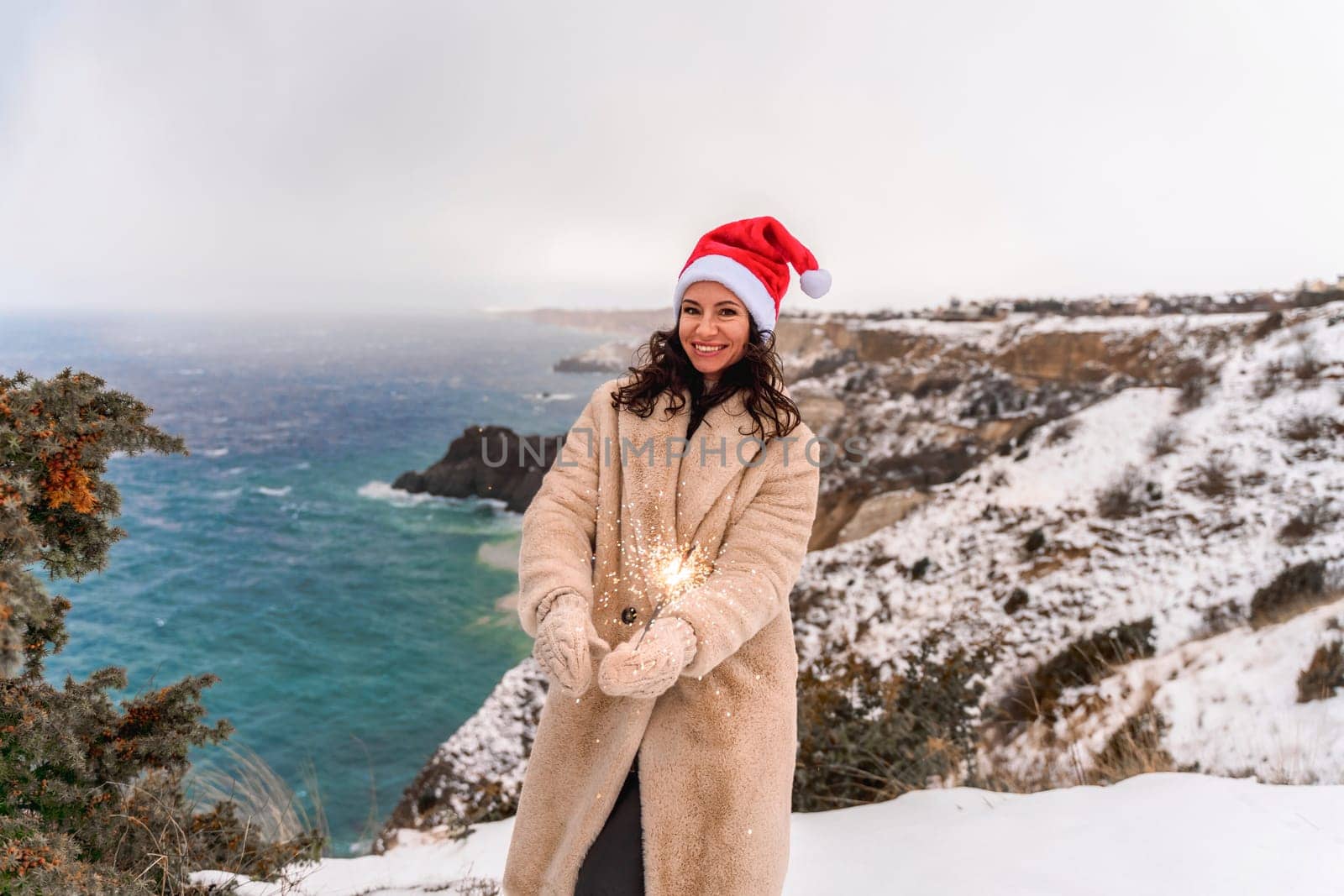 Outdoor winter portrait of happy smiling woman, light faux fur coat holding heart sparkler, posing against sea and snow background.