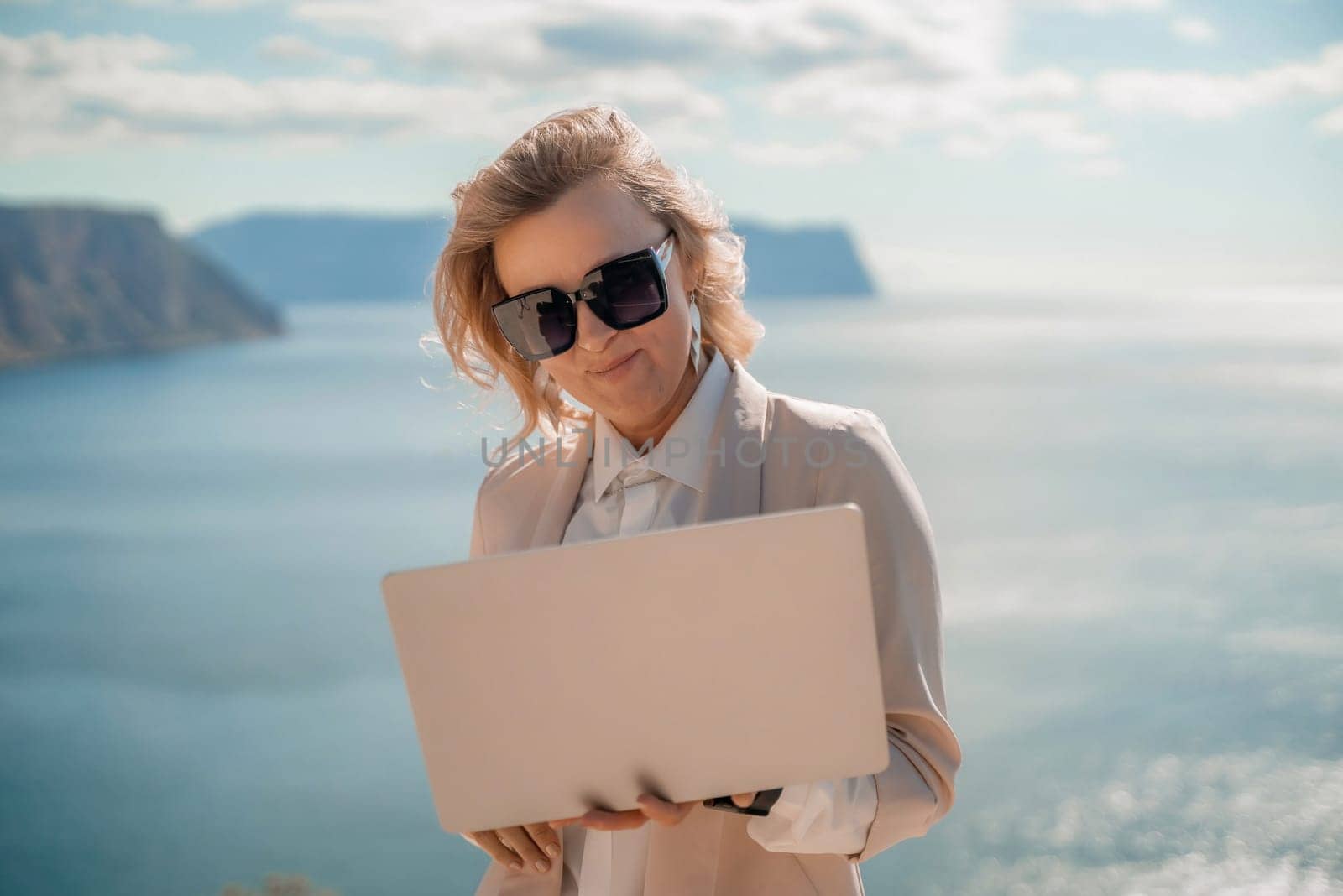 Freelance women sea. She is working on the computer. Good looking middle aged woman typing on a laptop keyboard outdoors with a beautiful sea view. The concept of remote work. by Matiunina