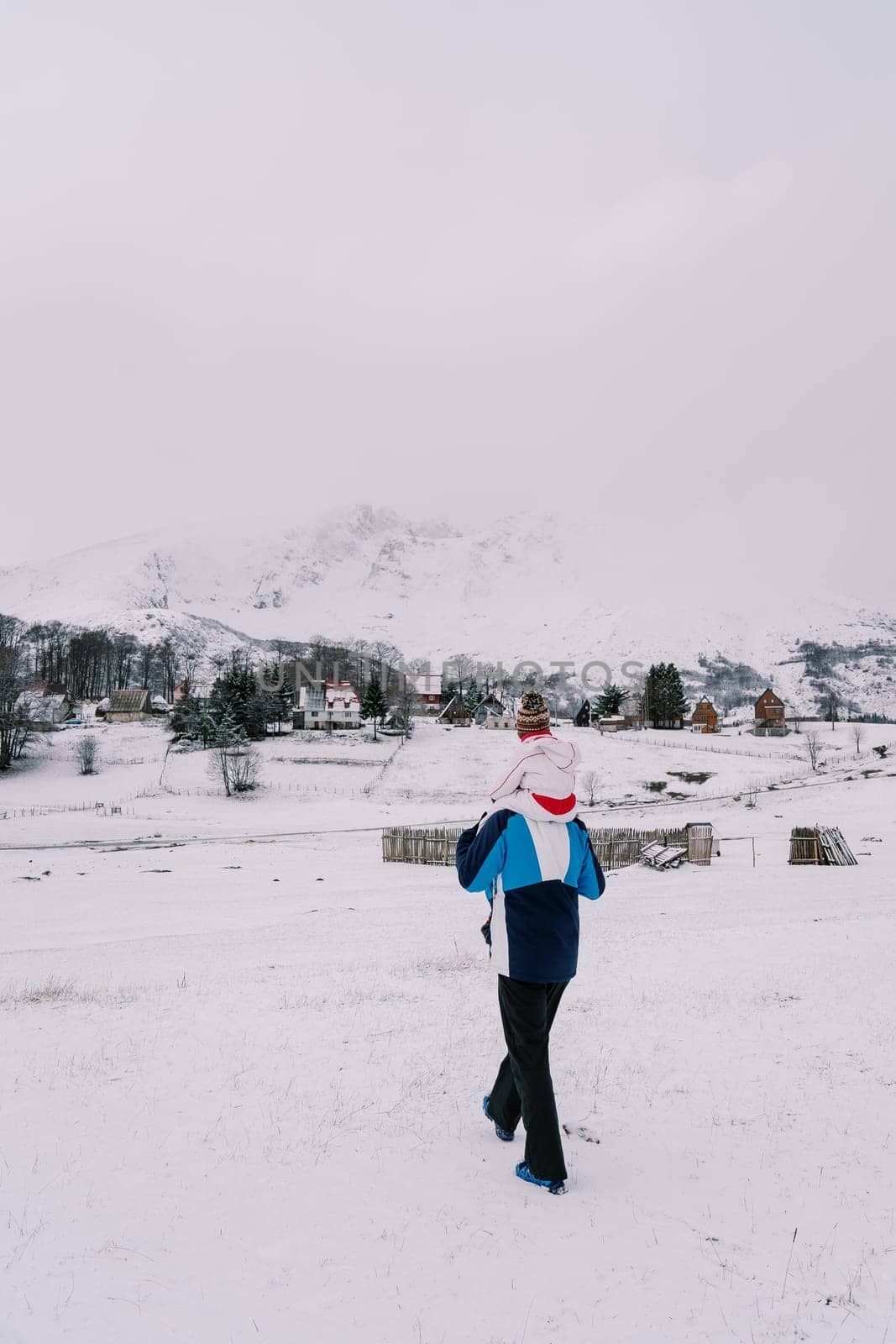 Father with a small child on his shoulders walks along a snow-covered hill towards a village at the foot of the mountains. Back view by Nadtochiy