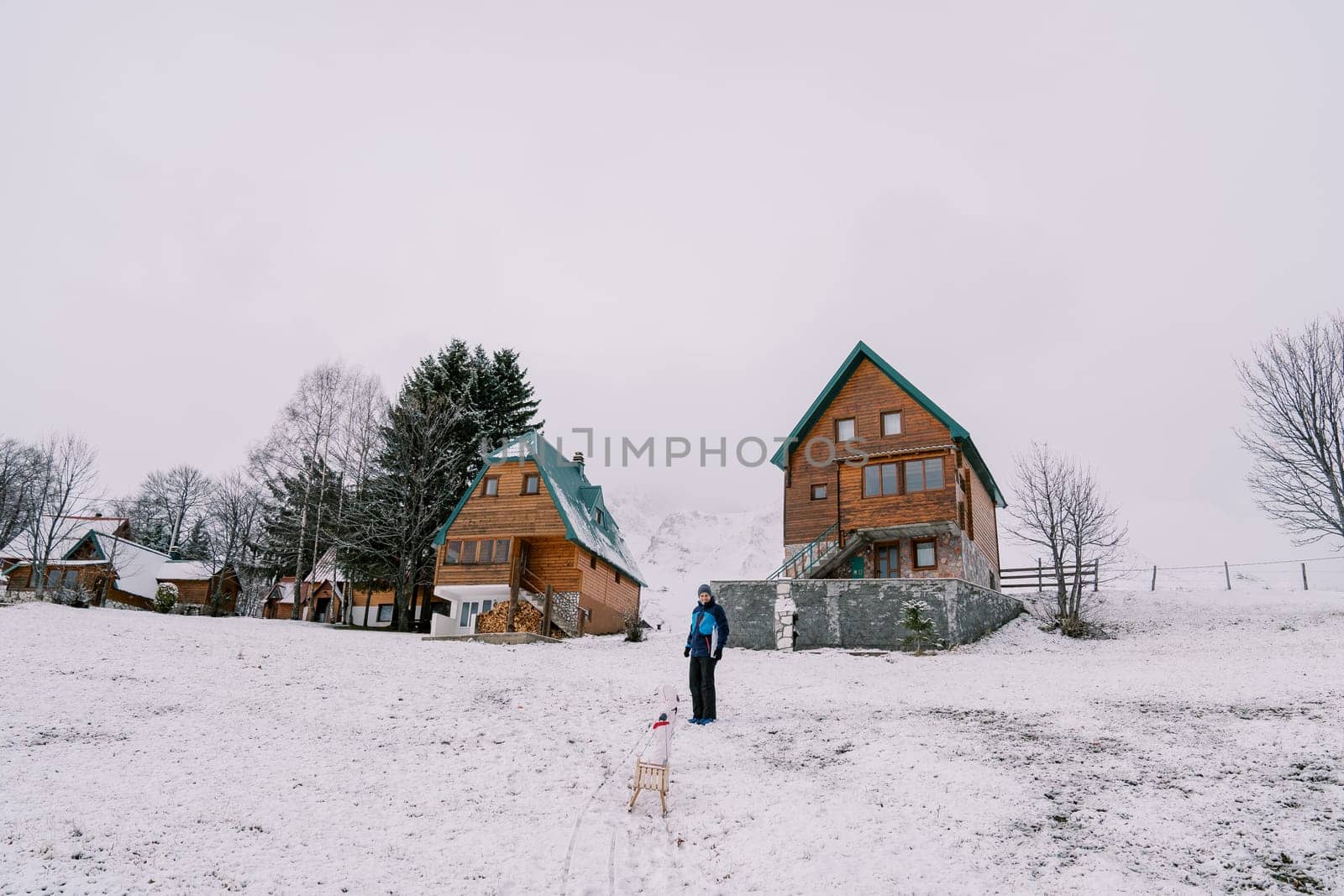 Dad stands near the house, looking at a small child climbing a hill with a sled. High quality photo