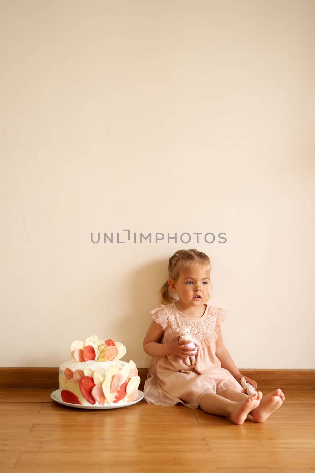 Little girl with a doll in her hand sits on the floor next to a birthday cake on a plate by Nadtochiy