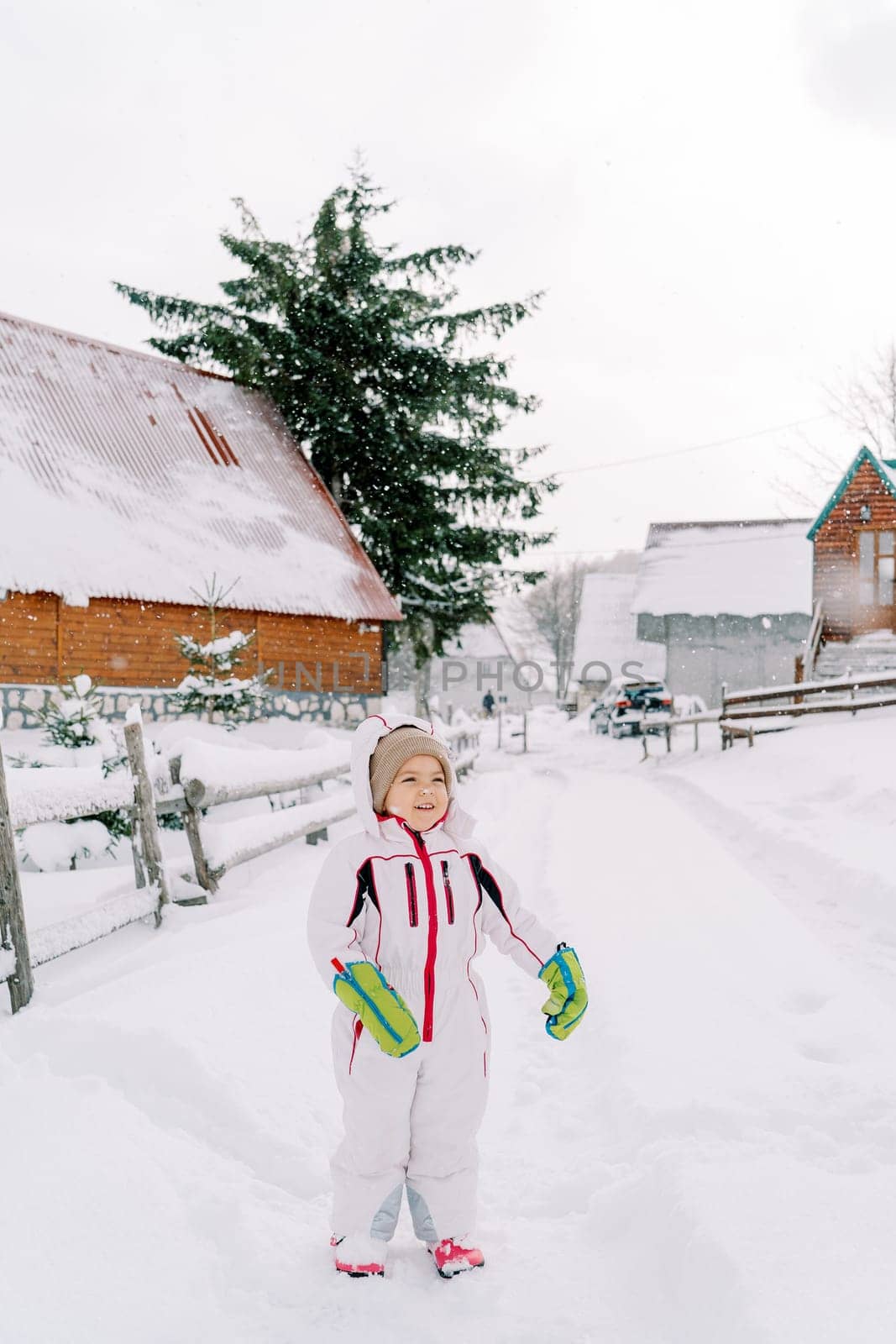 Little smiling girl stands on a snowy road near a fence in the village by Nadtochiy