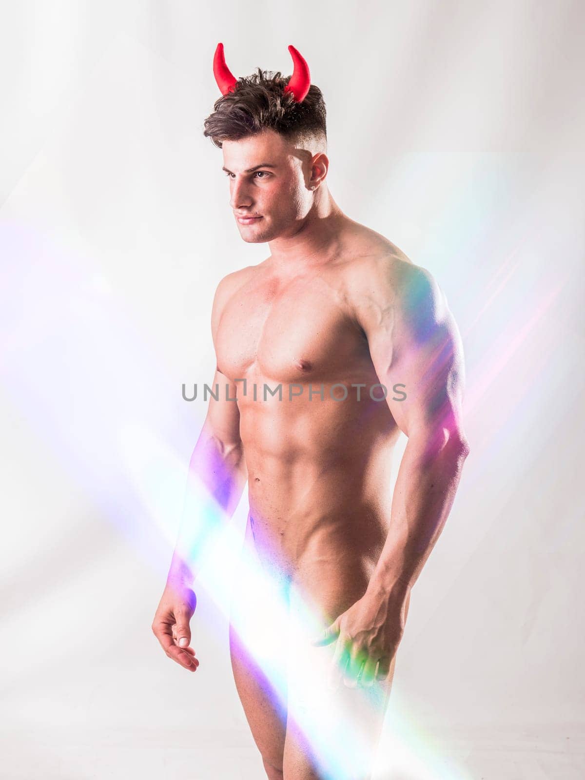 Muscular male bodybuilder totally nude, wearing devil costume horns on light background