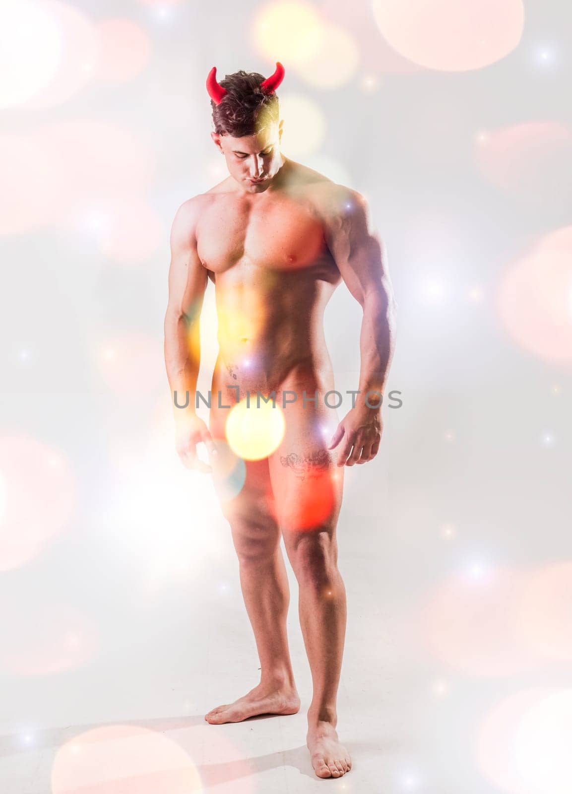 Muscular male bodybuilder totally nude, wearing devil costume horns on light background