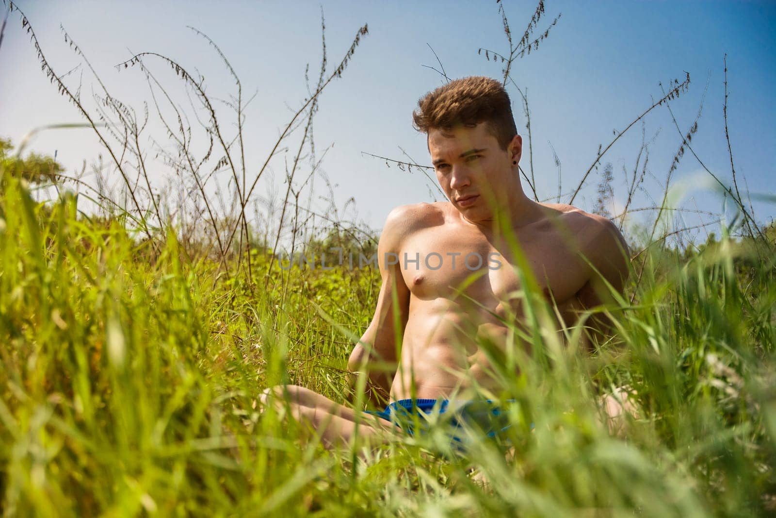 Handsome Muscular Shirtless Young Hunk Man Outdoor in Nature Sitting on Grass. Showing Healthy Muscle Body While Looking away