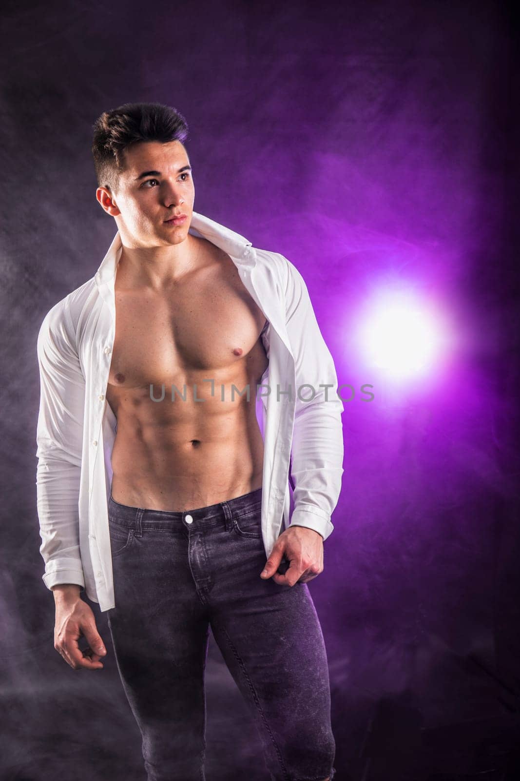 Handsome young muscular man shirtless wearing jeans, taking off leather jacket on naked muscle torso, on dark background in studio shot