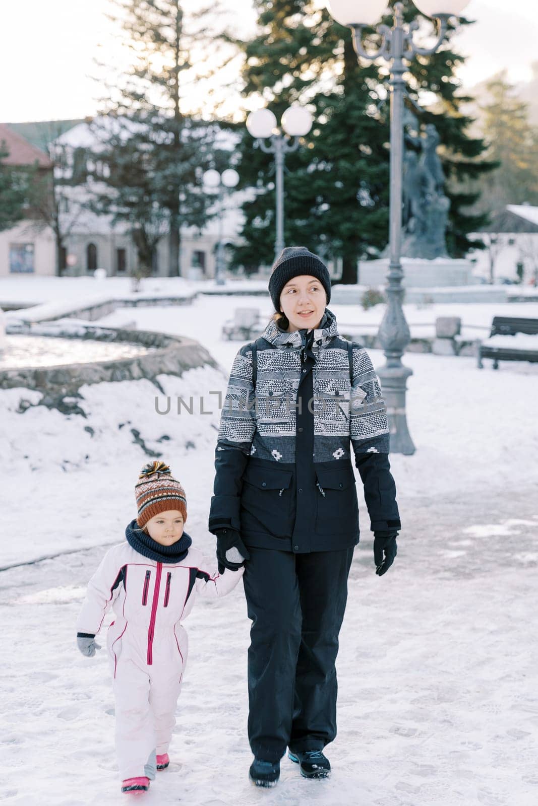 Mom and little girl walk holding hands through a snowy park by Nadtochiy
