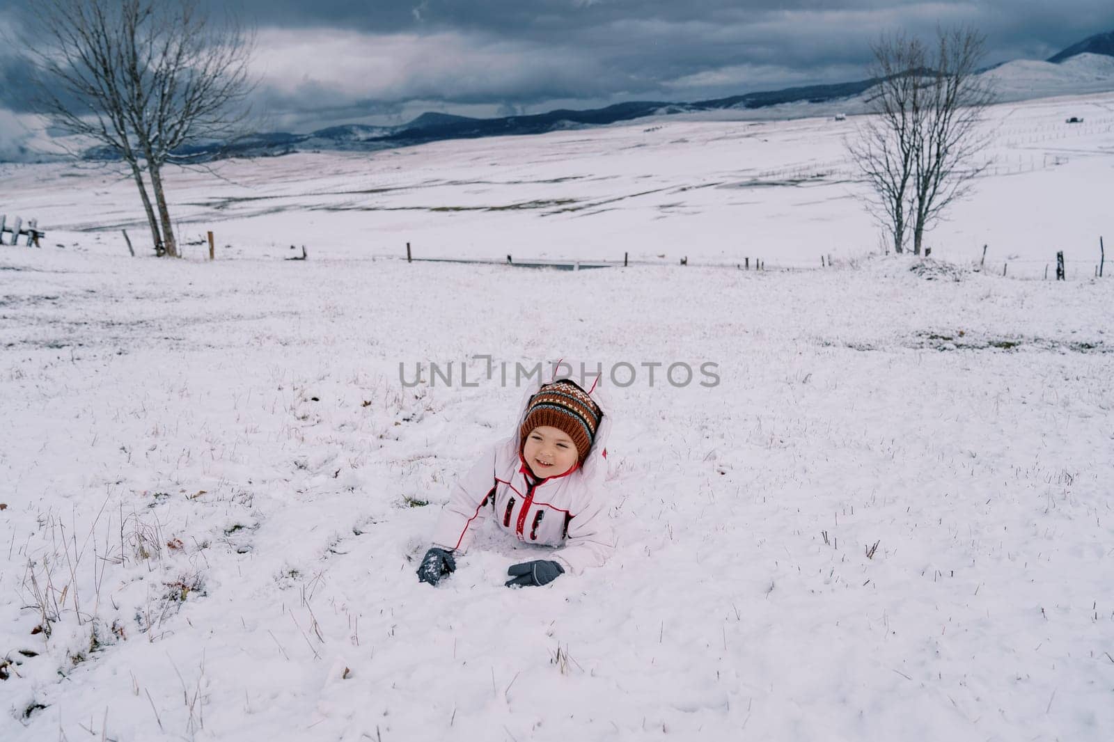 Little laughing girl lies on her stomach in the snow in a mountain valley. High quality photo