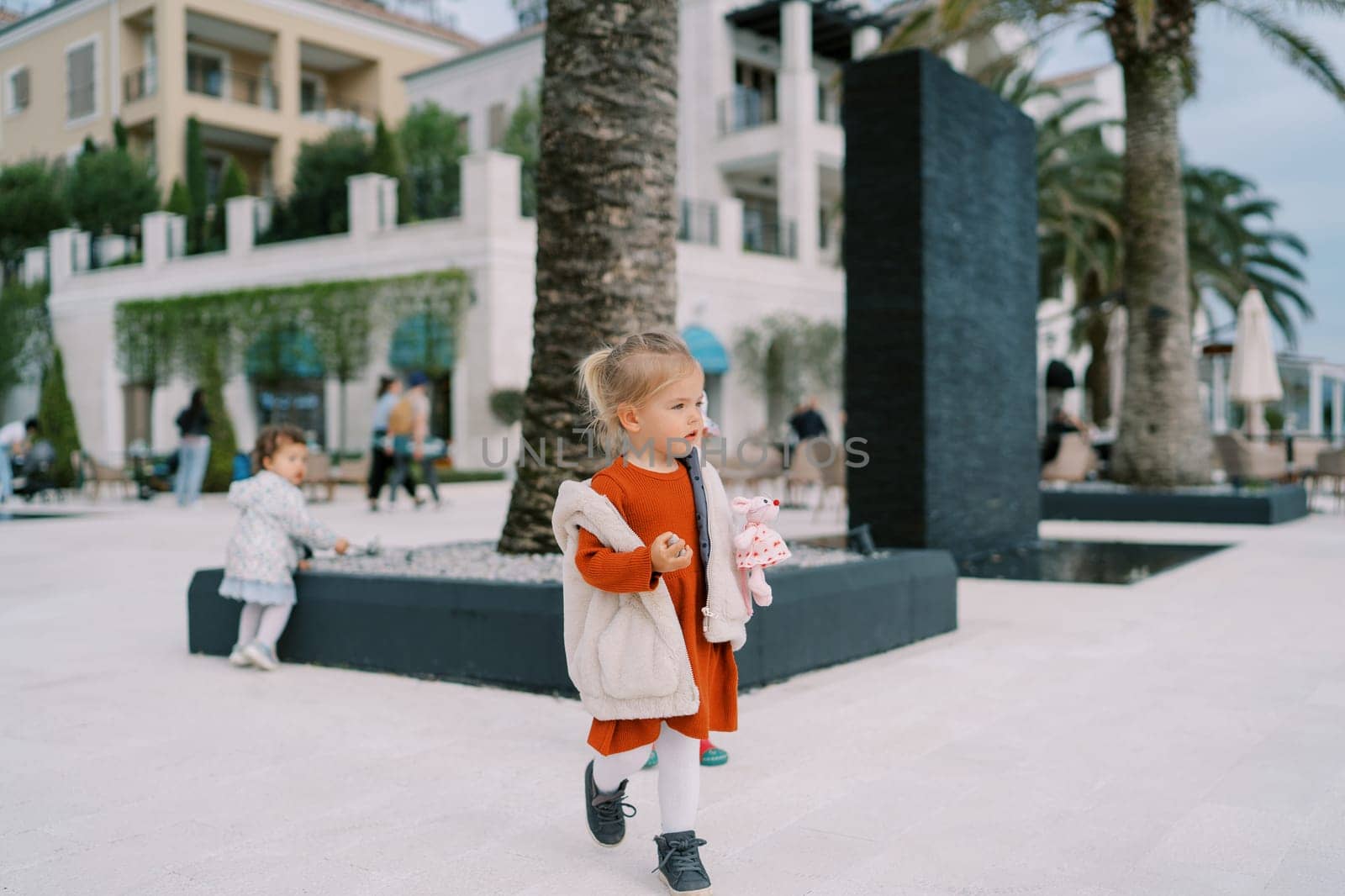 Little girl with a soft toy in her hand walks along the embankment of a resort town. High quality photo