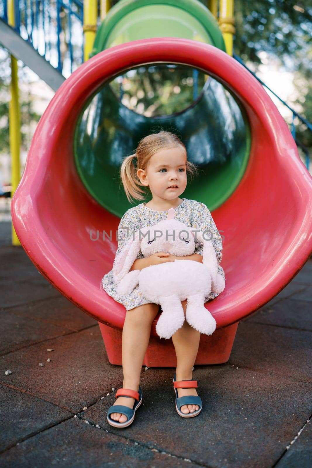Little girl with a toy pink rabbit sits on a tube slide and looks into the distance. High quality photo