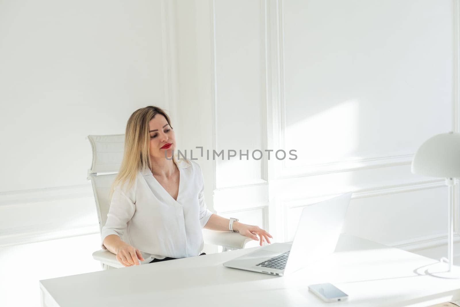 Delighted young blonde woman sitting at white table in blouse using laptop. High quality photo