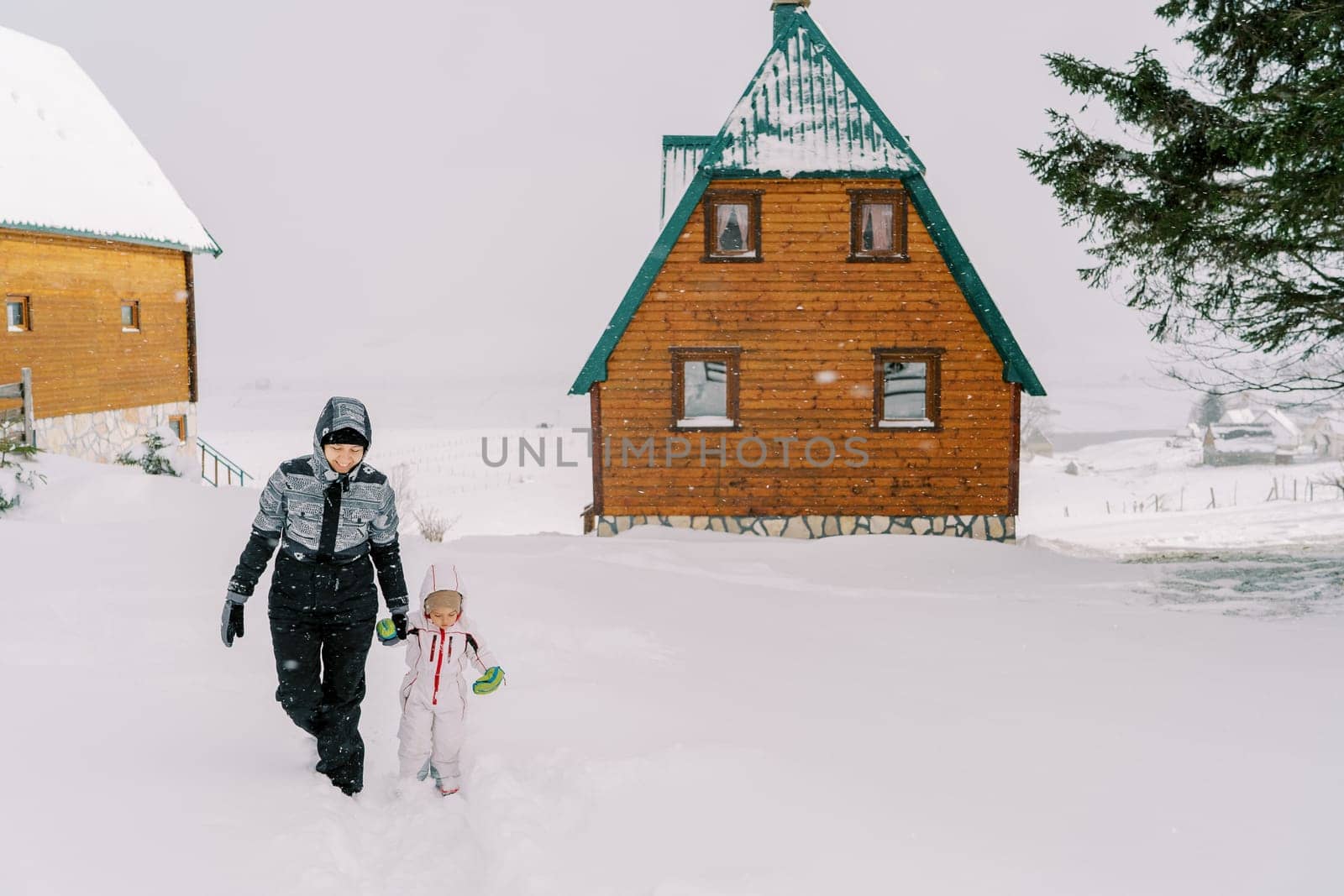 Mom and a little girl walk holding hands through the snowdrifts near wooden houses in the village. High quality photo