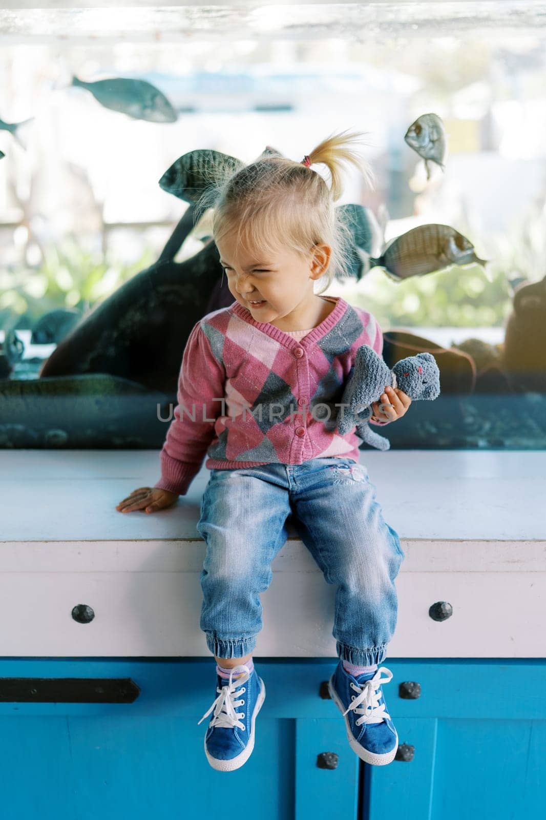 Little smiling girl with a soft toy sits on a table near an aquarium with fish. High quality photo