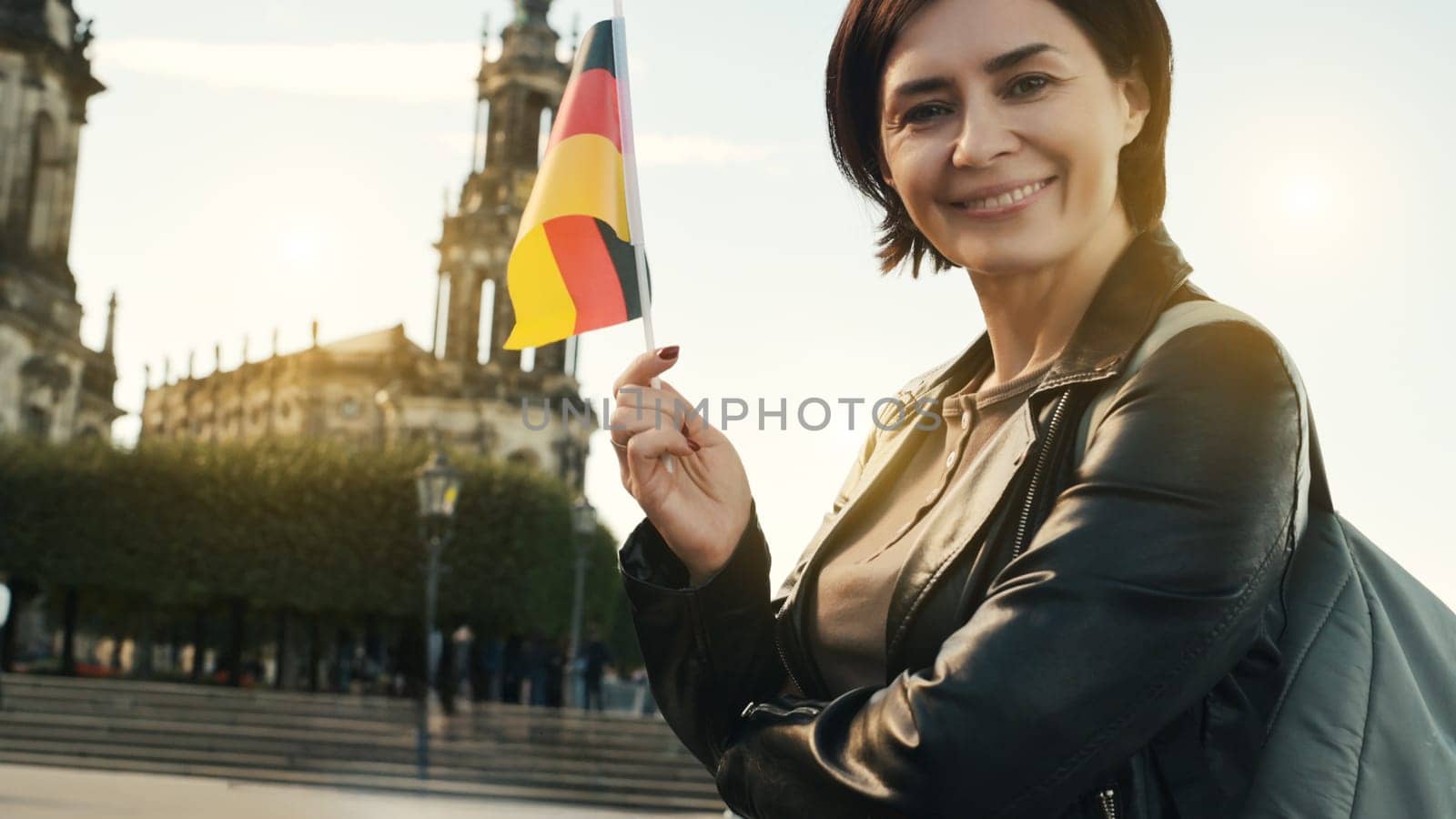 Young Woman Smiles With German Flag In Hand, Against Blurred City Backdrop In Autumn