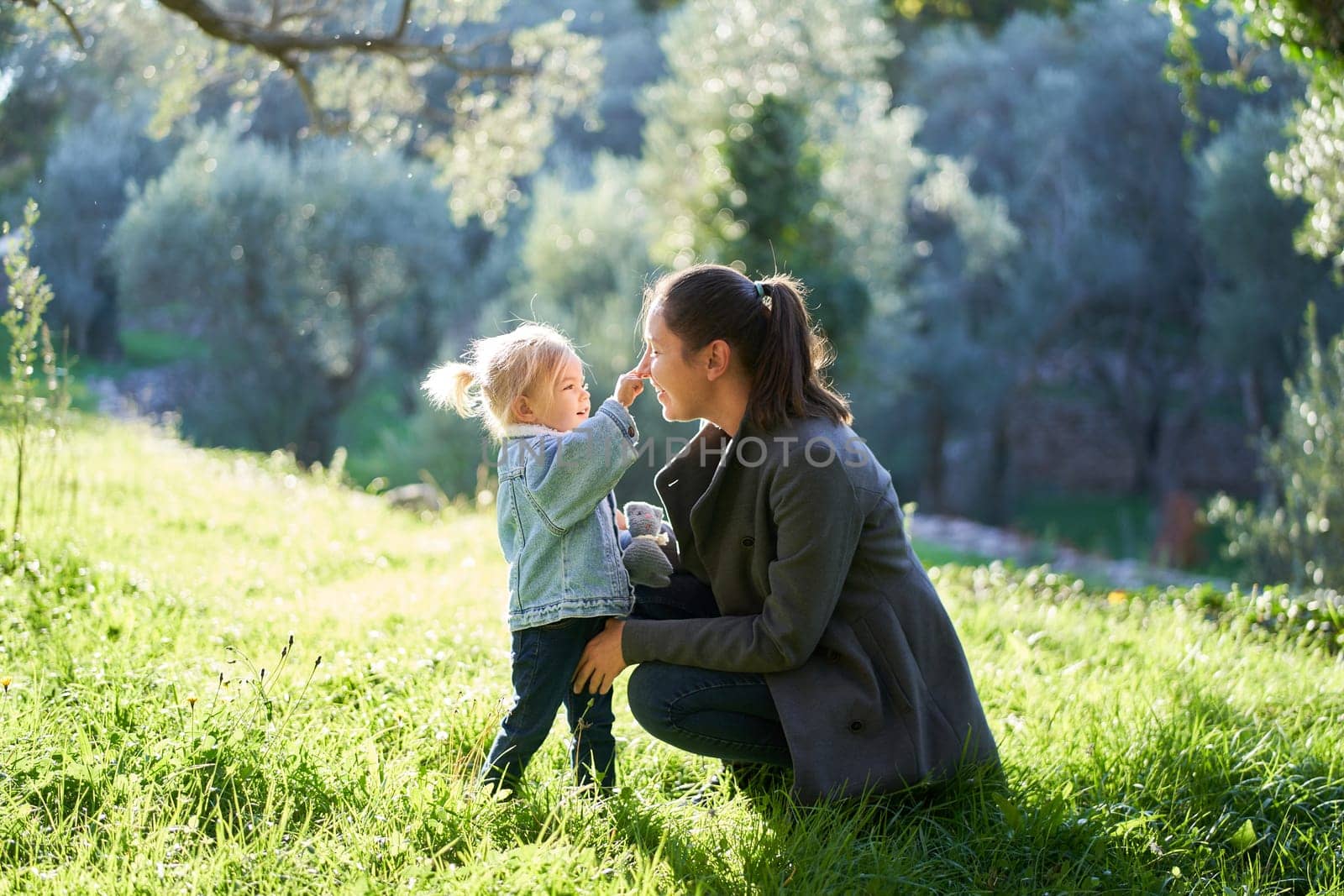 Little girl touches her mom nose while squatting on a green lawn by Nadtochiy