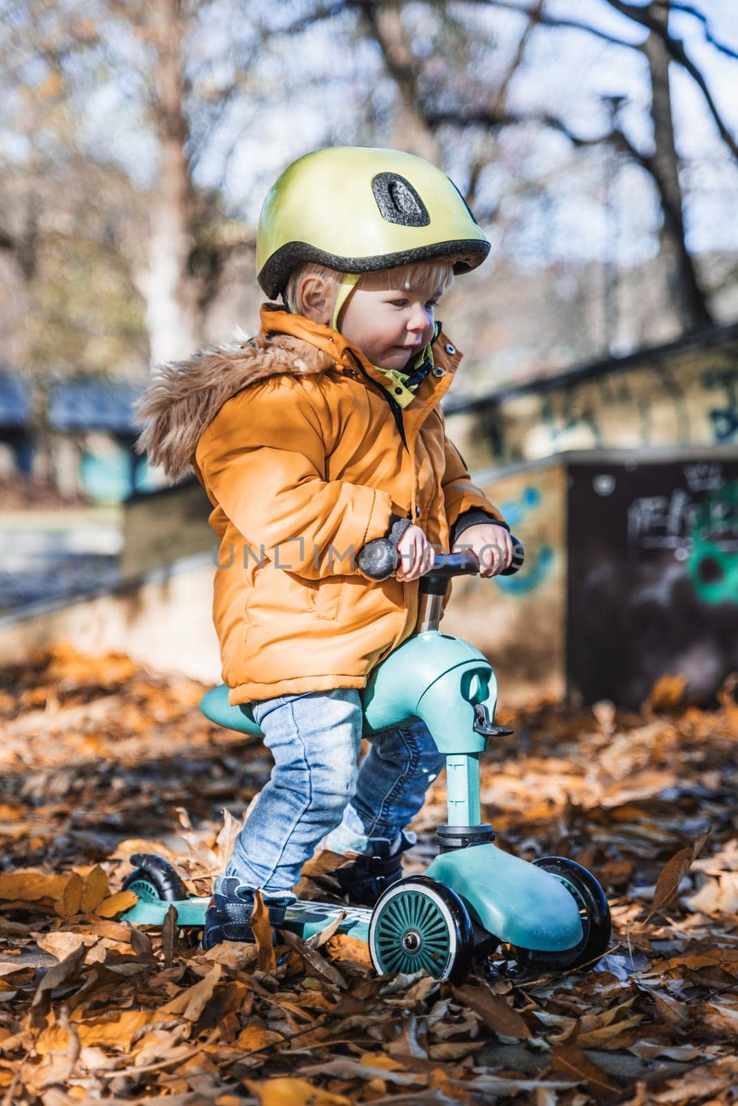 Adorable toddler boy wearing yellow protective helmet riding baby scooter outdoors on autumn day. Kid training balance on mini bike in city park. Fun autumn outdoor activity for small kids. by kasto