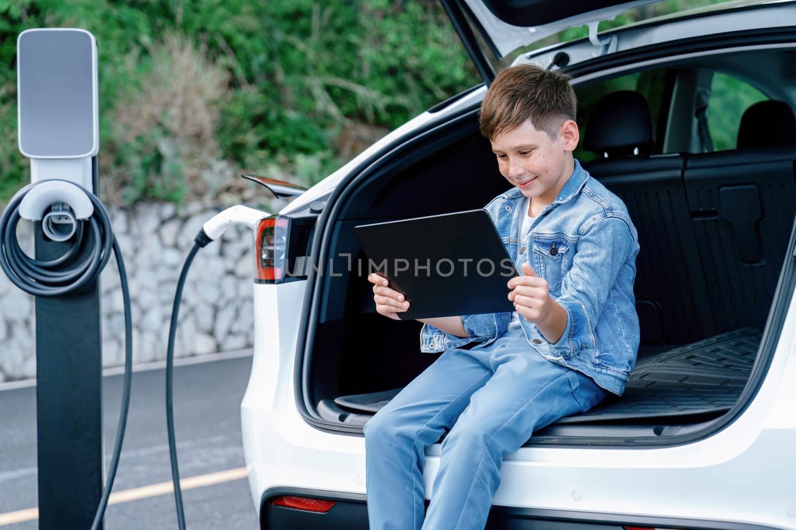 Little boy sitting on car trunk, using tablet while recharging eco-friendly car from EV charging station. EV car road trip travel as alternative vehicle using sustainable energy concept. Perpetual