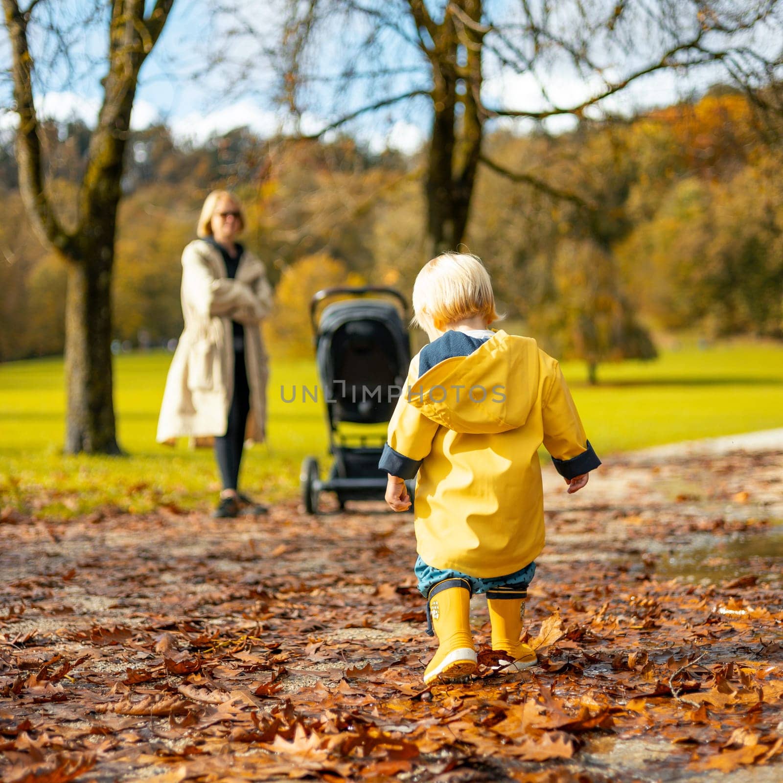 Sun always shines after the rain. Small bond infant boy wearing yellow rubber boots and yellow waterproof raincoat walking in puddles and autumn leaves in city park on sunny rainy day