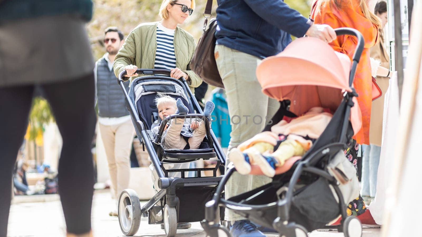 Mother walking and pushing his infant baby boy child in stroller in crowd of people wisiting sunday flea market in Malaga, Spain. by kasto