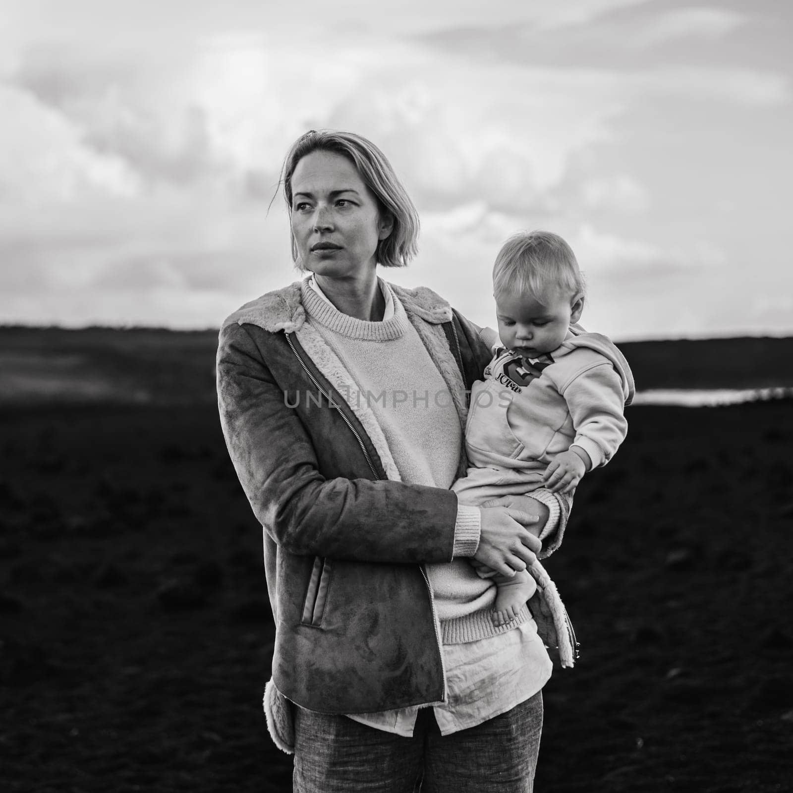 Mother enjoying winter vacations playing with his infant baby boy son on black sandy volcanic beach of Janubio on Lanzarote island, Spain on windy overcast day. Family travel vacations concept. by kasto