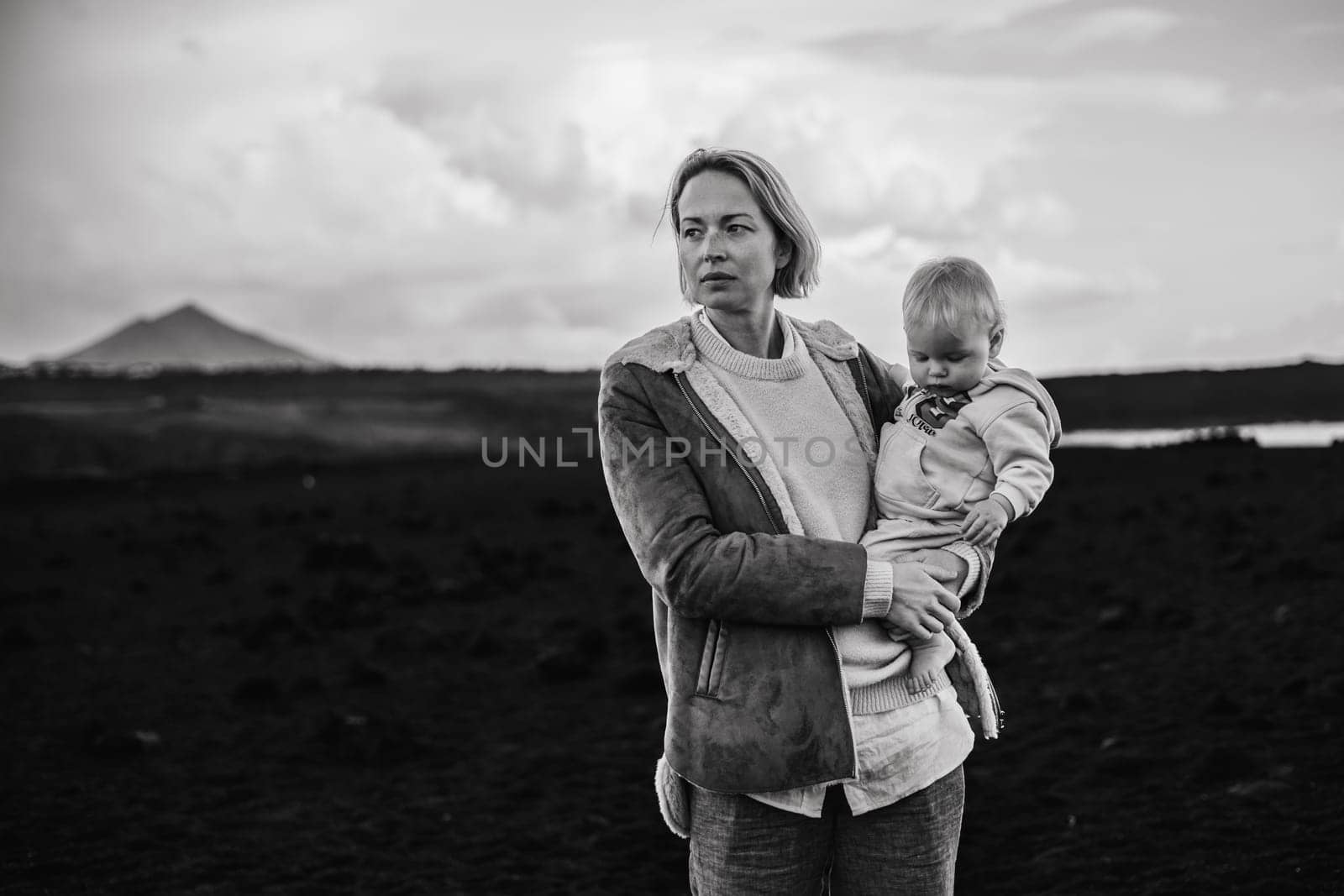 Mother enjoying winter vacations playing with his infant baby boy son on black sandy volcanic beach of Janubio on Lanzarote island, Spain on windy overcast day. Family travel vacations concept. by kasto