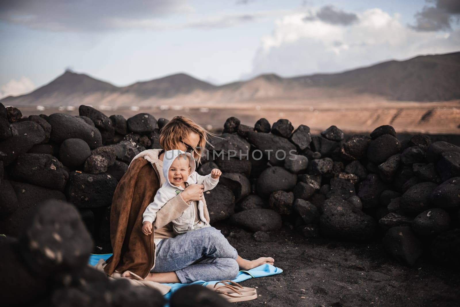 Mother enjoying winter vacations playing with his infant baby boy son on black sandy volcanic beach of Janubio on Lanzarote island, Spain on windy overcast day. Family travel vacations concept. by kasto