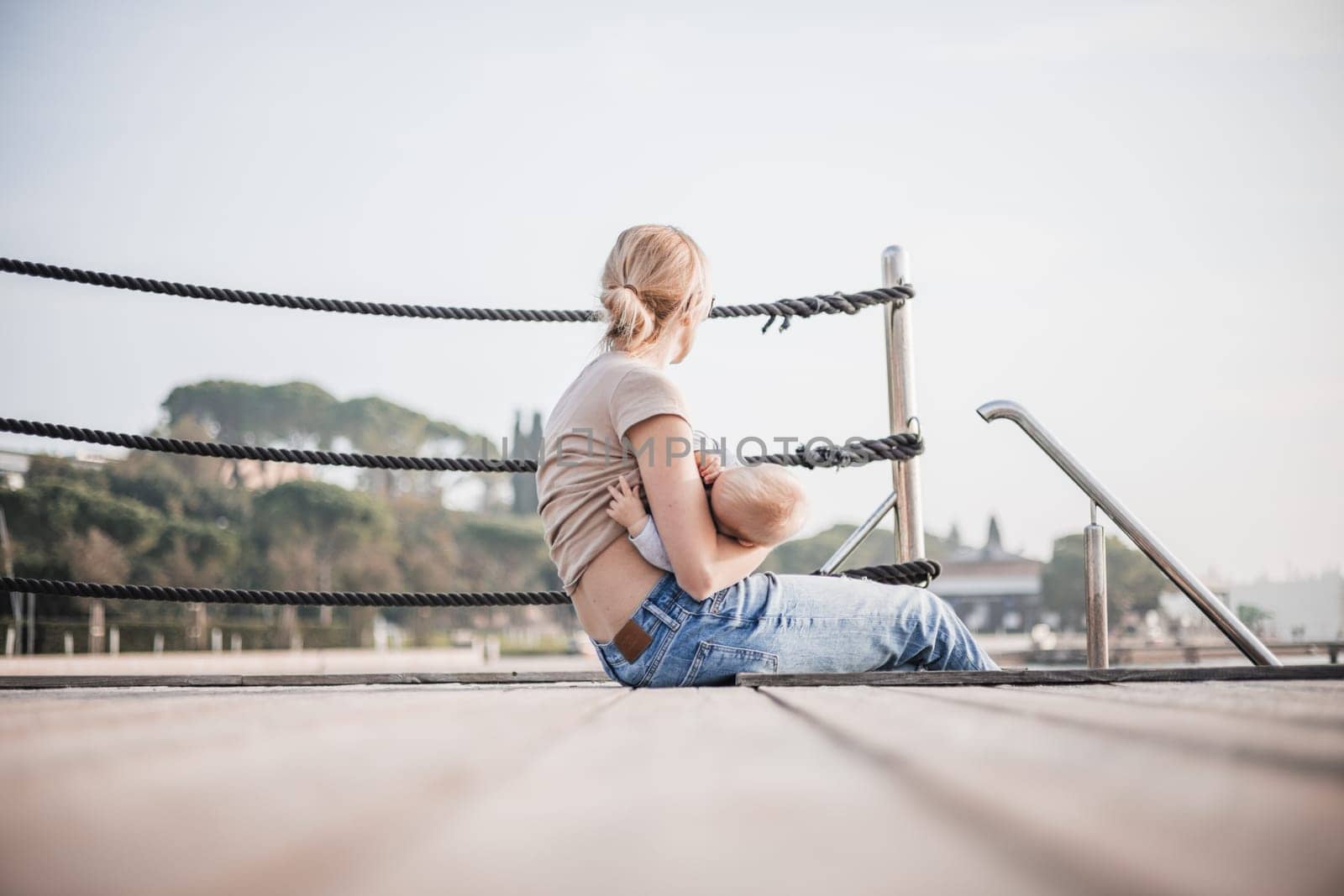 Mother breast feeding her little baby boy infant child on old wooden pier by the sea on nice warm spring day enjoing nature and warm breeze outdoors