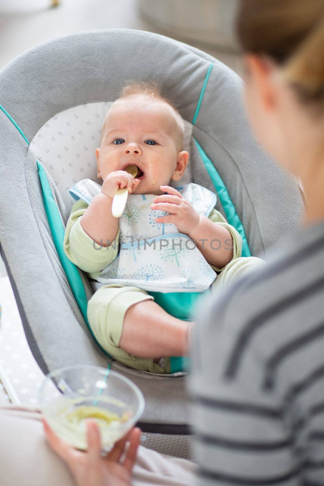 Mother spoon feeding her baby boy child in baby chair with fruit puree. Baby solid food introduction concept