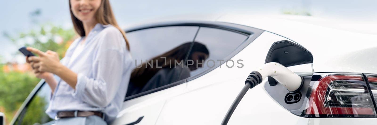 Young woman using smartphone online banking application to pay for electric car battery charging from EV charging station during vacation road trip at national park or summer forest. Panorama Exalt