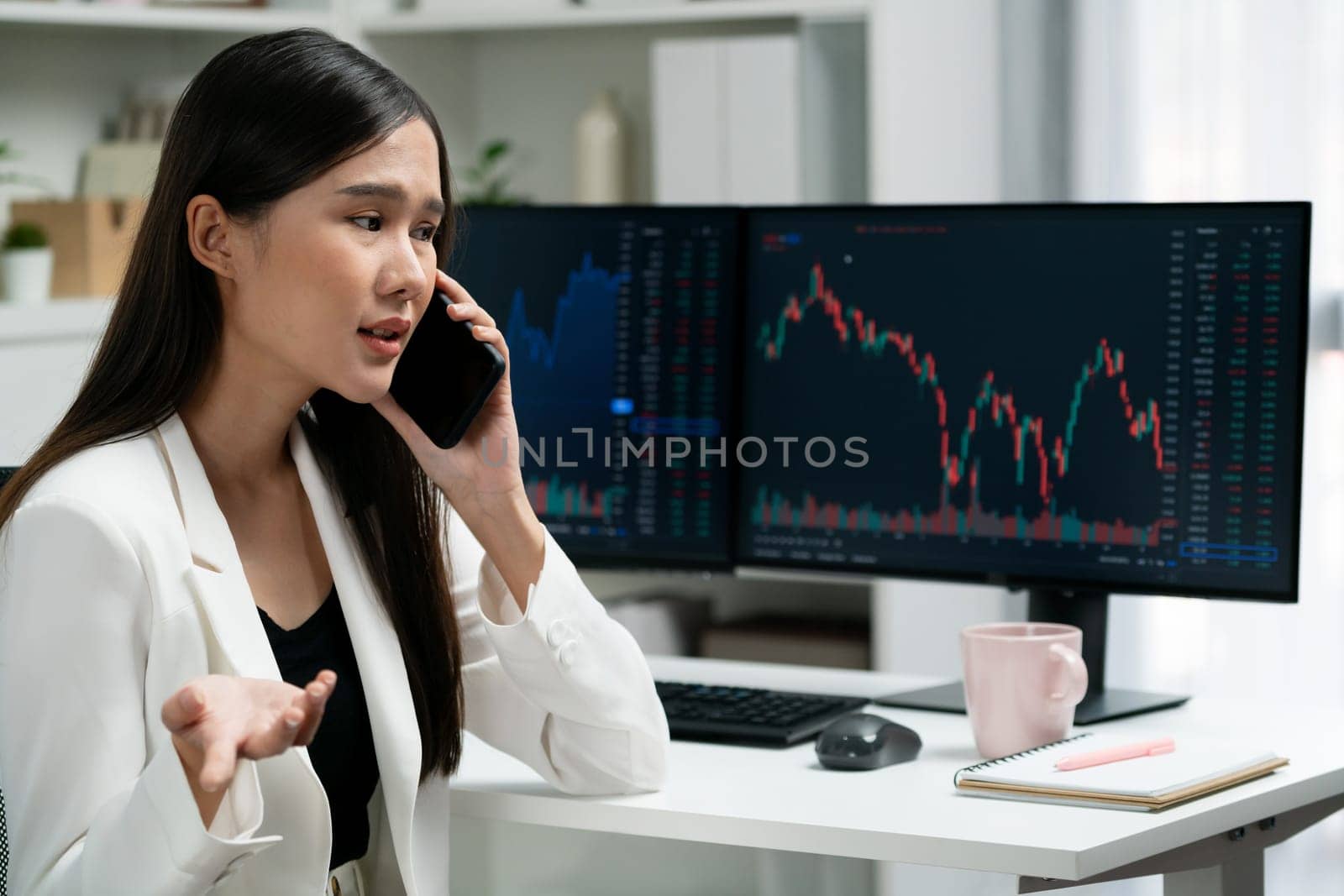 Stressful young beautiful Asian businesswoman calling to broker on smartphone, analyze possible risk to profit or loss in dynamic exchange stock market graph on pc screen at modern office. Stratagem.