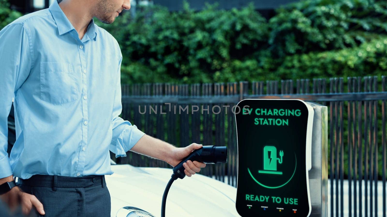 Businessman pull and hold EV charger plug form electric car charging station at city car park area background. Futuristic clean sustainable energy and EV car technological advancement.Peruse