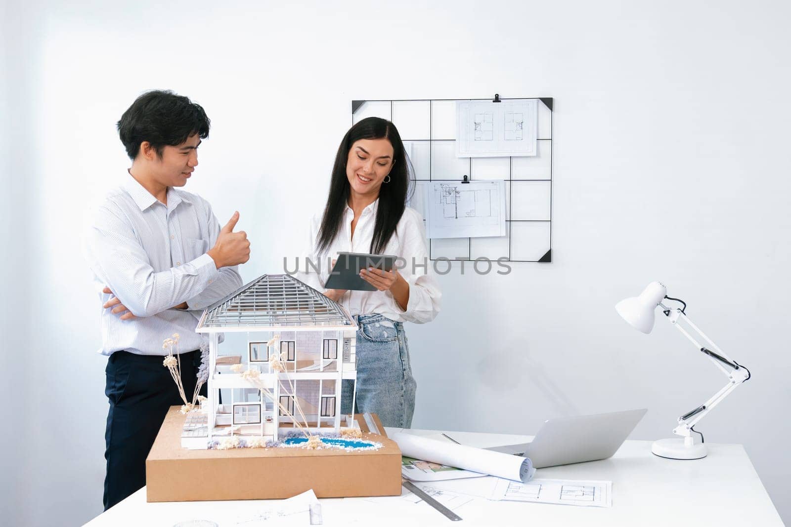 Beautiful young caucasian project manager tells asian engineer about building detail by using tablet while engineer measuring house model on meeting table with blueprint scatter around. Immaculate