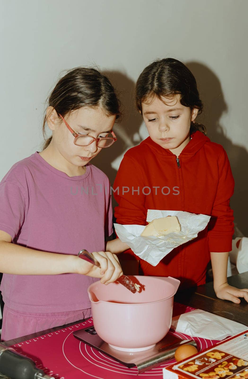 Two beautiful Caucasian brunette girls are standing at a table with food and one is cutting a piece of butter into a bowl for baking cookies, standing at the table in the kitchen during the day, close-up side view. Step-by-step instructions. Step 2.