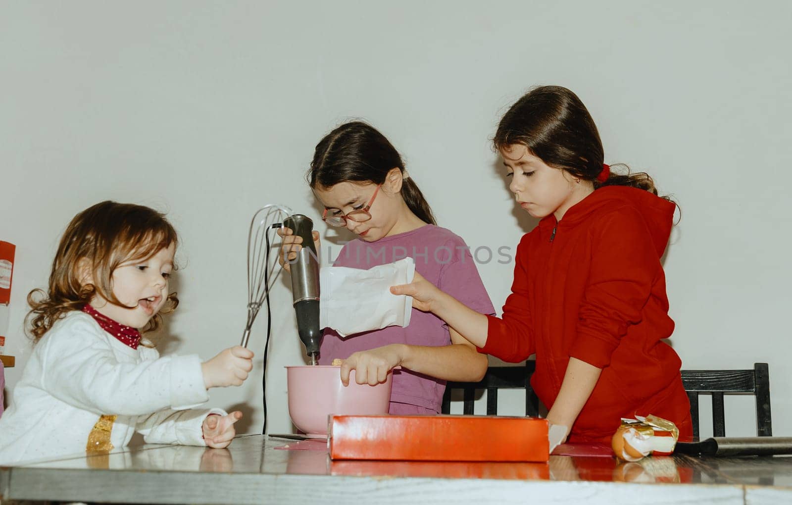 Three beautiful Caucasian brunette girls stand at the table and one pours the prepared flour mixture into a bowl for baking cookies, and the other beats it with a mixer, the third plays with a whisk, close-up side view. Step-by-step instructions. Step 6.